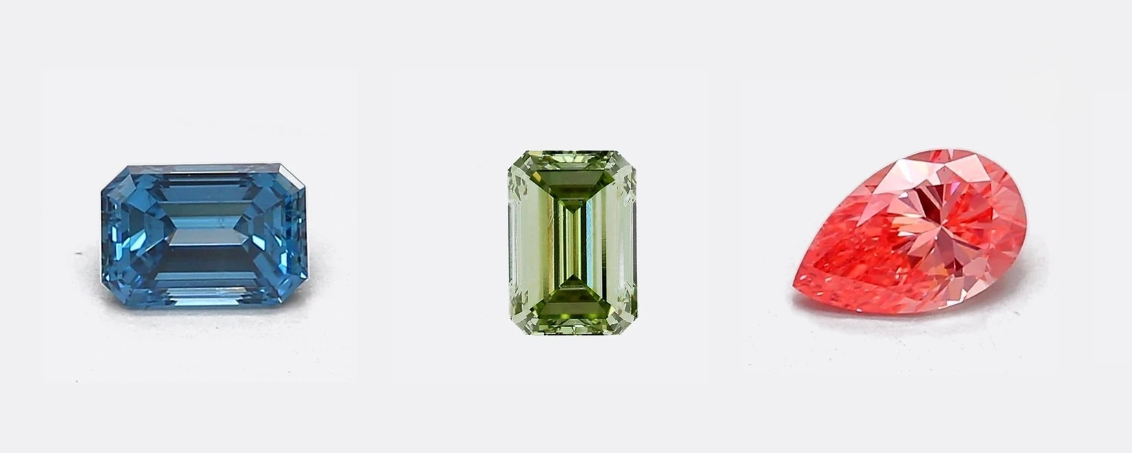Three loose fancy color diamonds side by side on a neutral gray background: a blue emerald-cut, a green emerald-cut, and a pink pear-cut diamond.